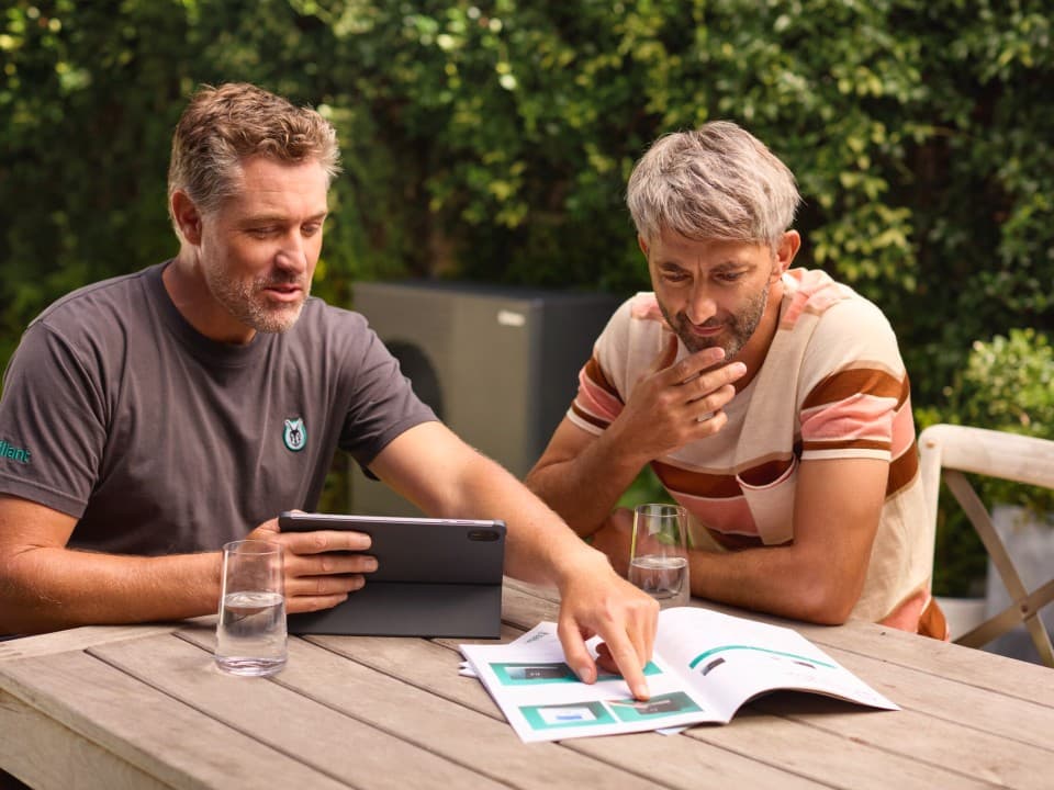 Zwei Männer in einem Garten, die auf einen Computer schauen, mit einer Wärmepumpe im Hintergrund.