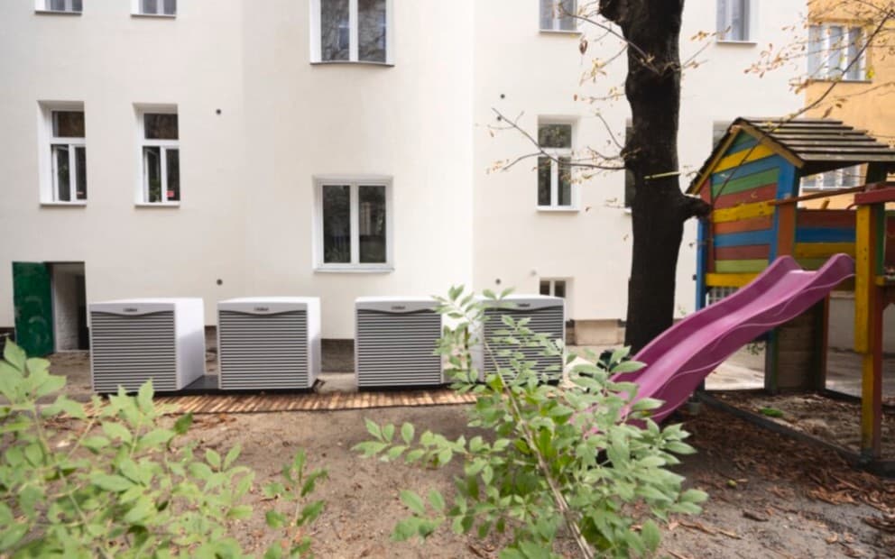Vier Wärmepumpen Außeneinheiten vor einem weißen Gebäude mit einem Spielplatz daneben.